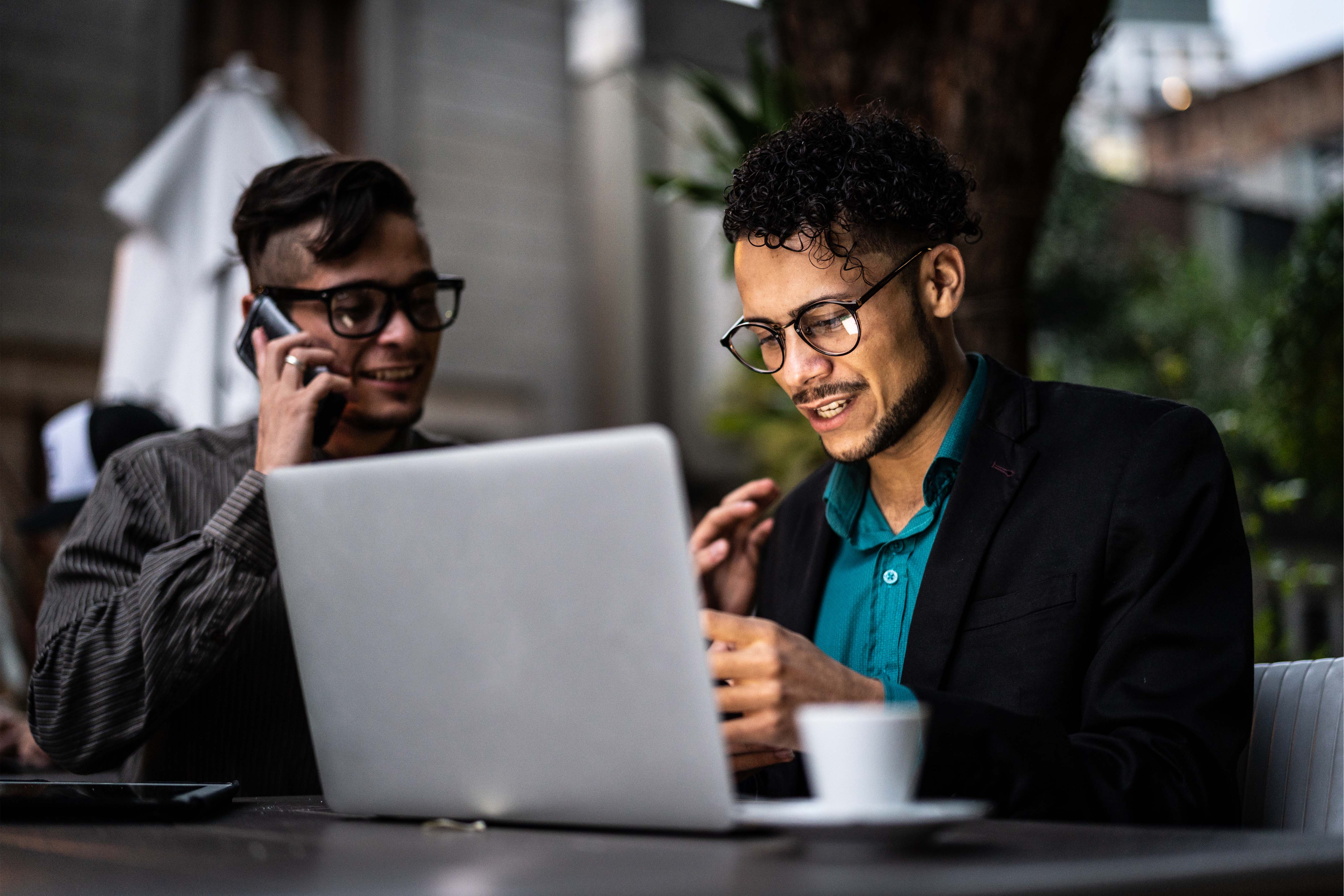 Two colleagues collaborate on a laptop at an outdoor café, one on a phone call, both smiling