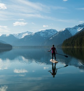 A woman paddle boards on a calm lake surrounded by mountains