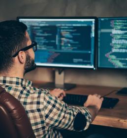 A bearded man in glasses sits in a leather chair at a wooden desk, coding across two monitors