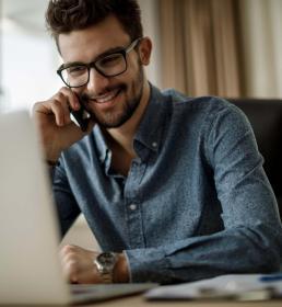 A smiling young businessman sits at his computer and speaks on the phone