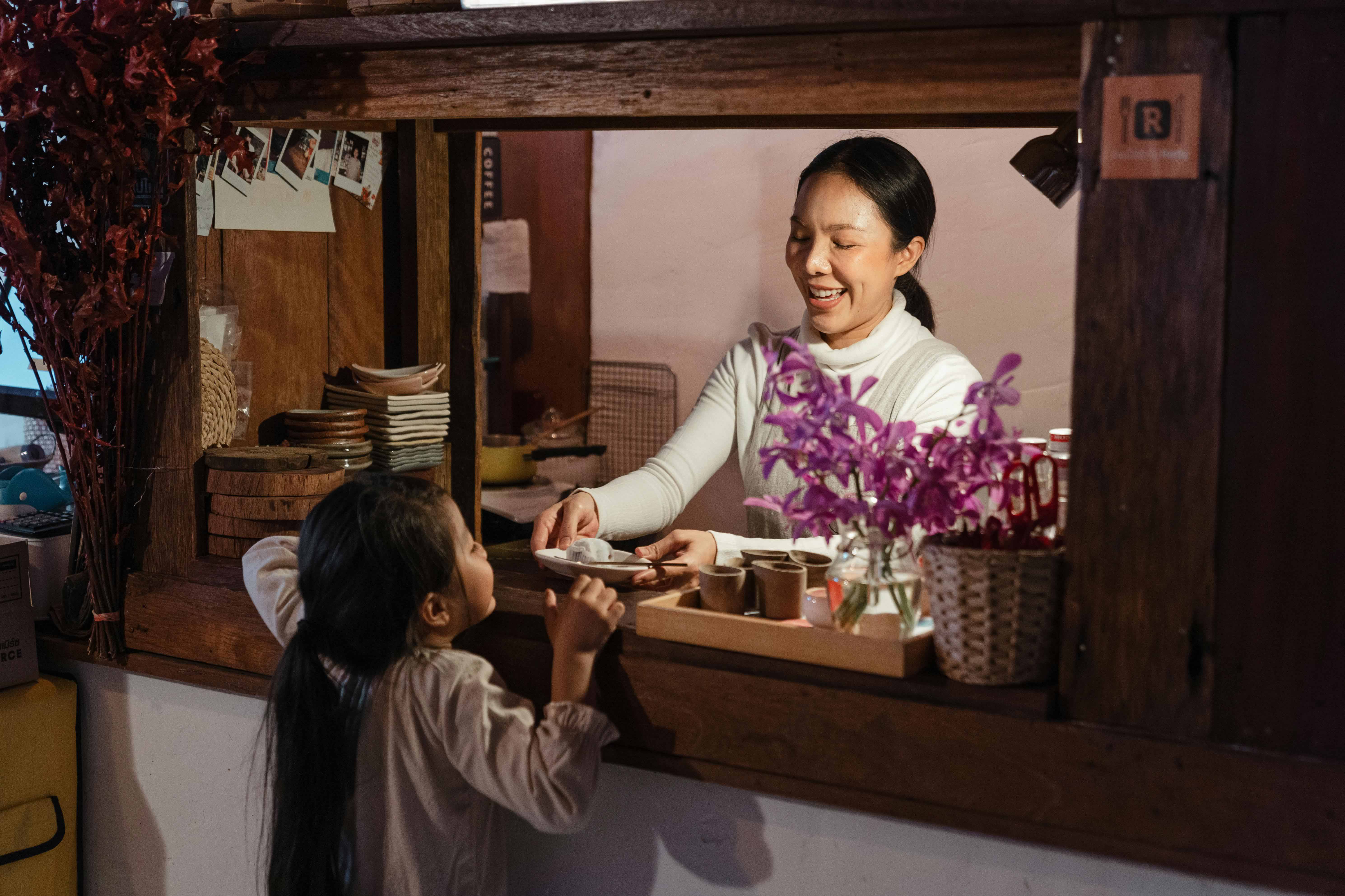 Smiling woman in a white top handing a plate to a young girl over a wooden café counter in Thailand