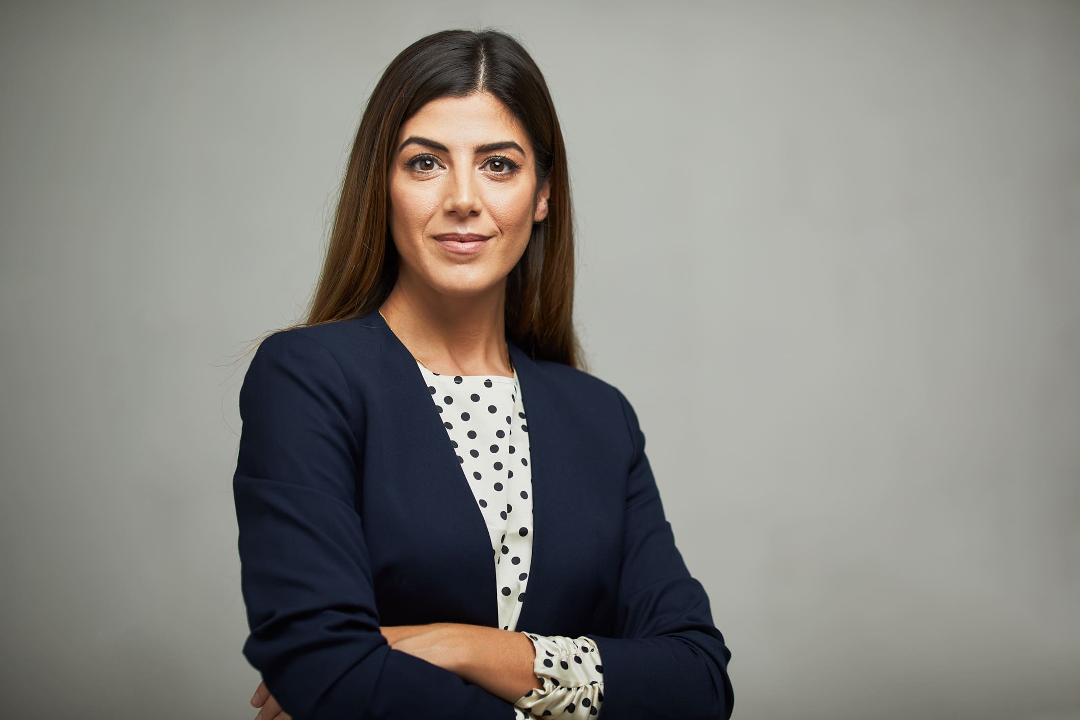 A woman in business attire stands against a plain background and smiles into the camera