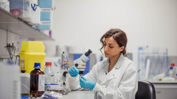 A female researcher or biochemist, sitting in a lab wearing a lab coat and blue plastic gloves, analyses a specimen in a test tube