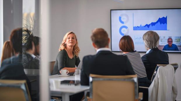 A blonde businesswoman listens to her colleague during a video conference call in a busy boardroom