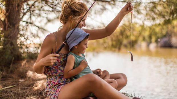 A woman and her young son are fishing by the side of a sunny lake. She holds up a small fish on the line as he smiles happily