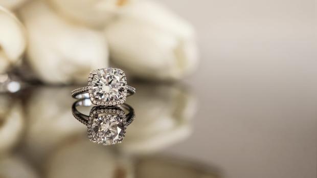 A diamond ring sitting on a reflecting surface in front of some white roses