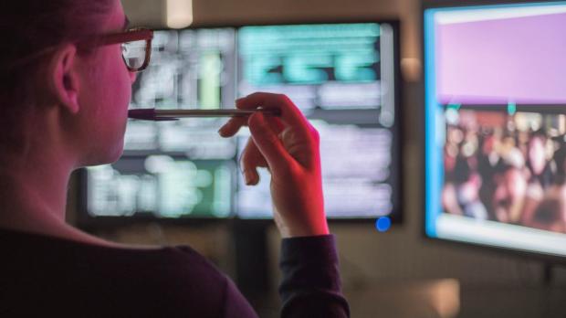 A computer technician sits looking at two monitor screens. She is illuminated by the red light from her screens