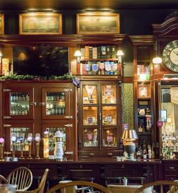 Wooden bar with glass cabinets, beer taps, a large clock, shelves of bottles, and dim lighting.