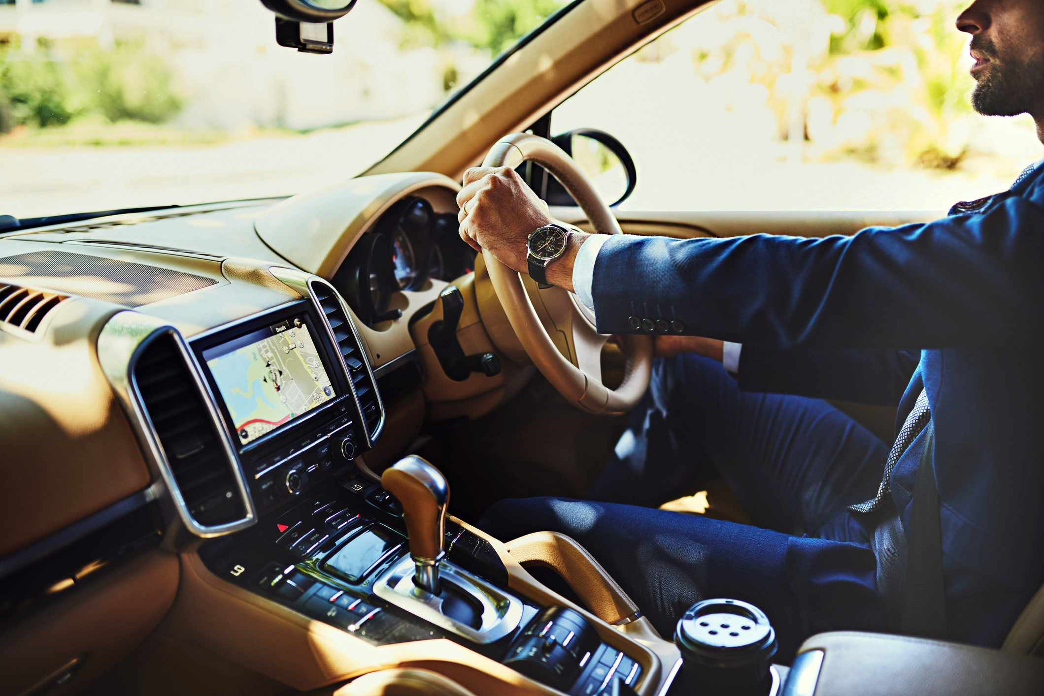 A handsome man in a tailored suit sits in an expensive car with his hands on the steering wheel