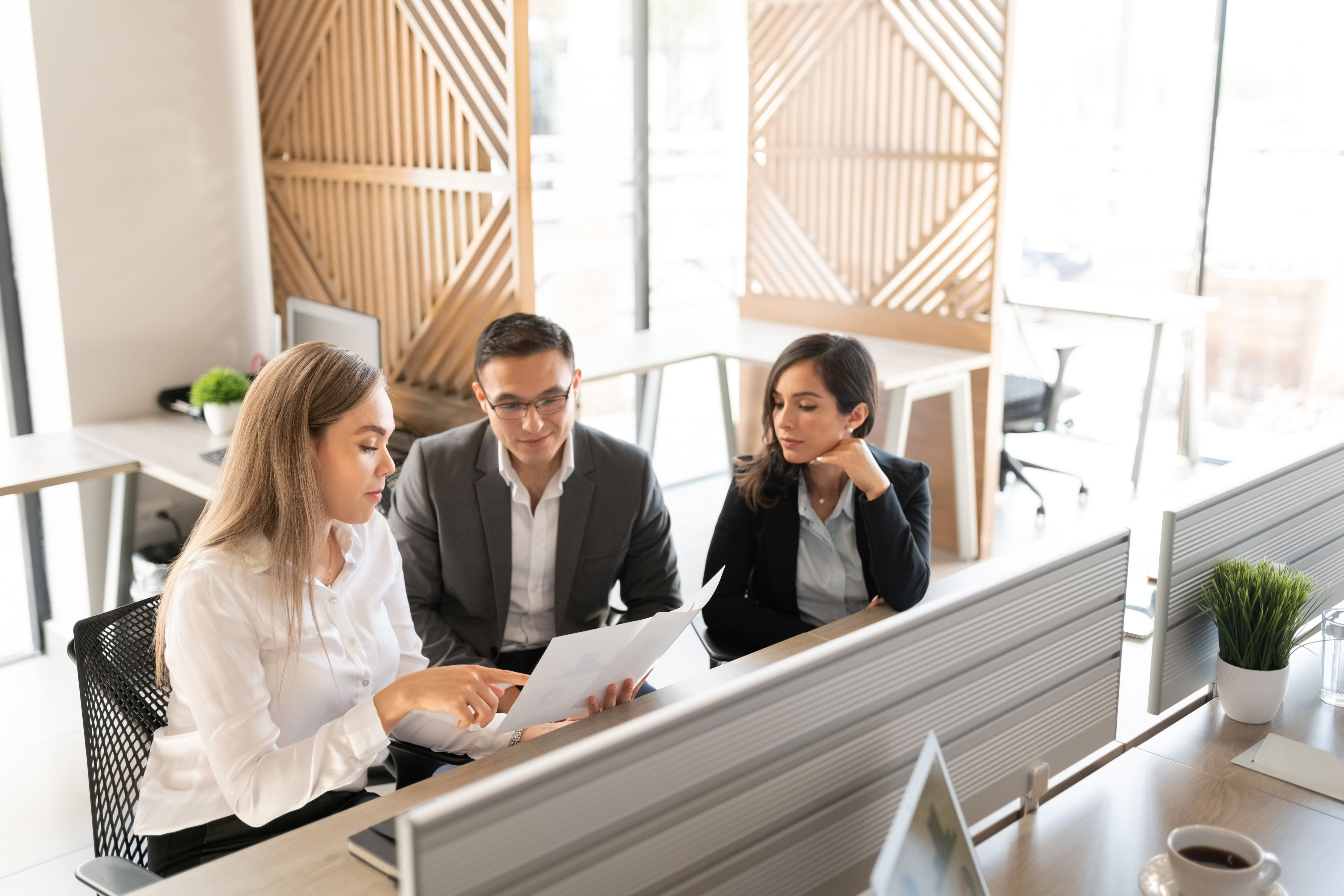 Three business professionals in a modern office discussing and reviewing documents at a workstation.