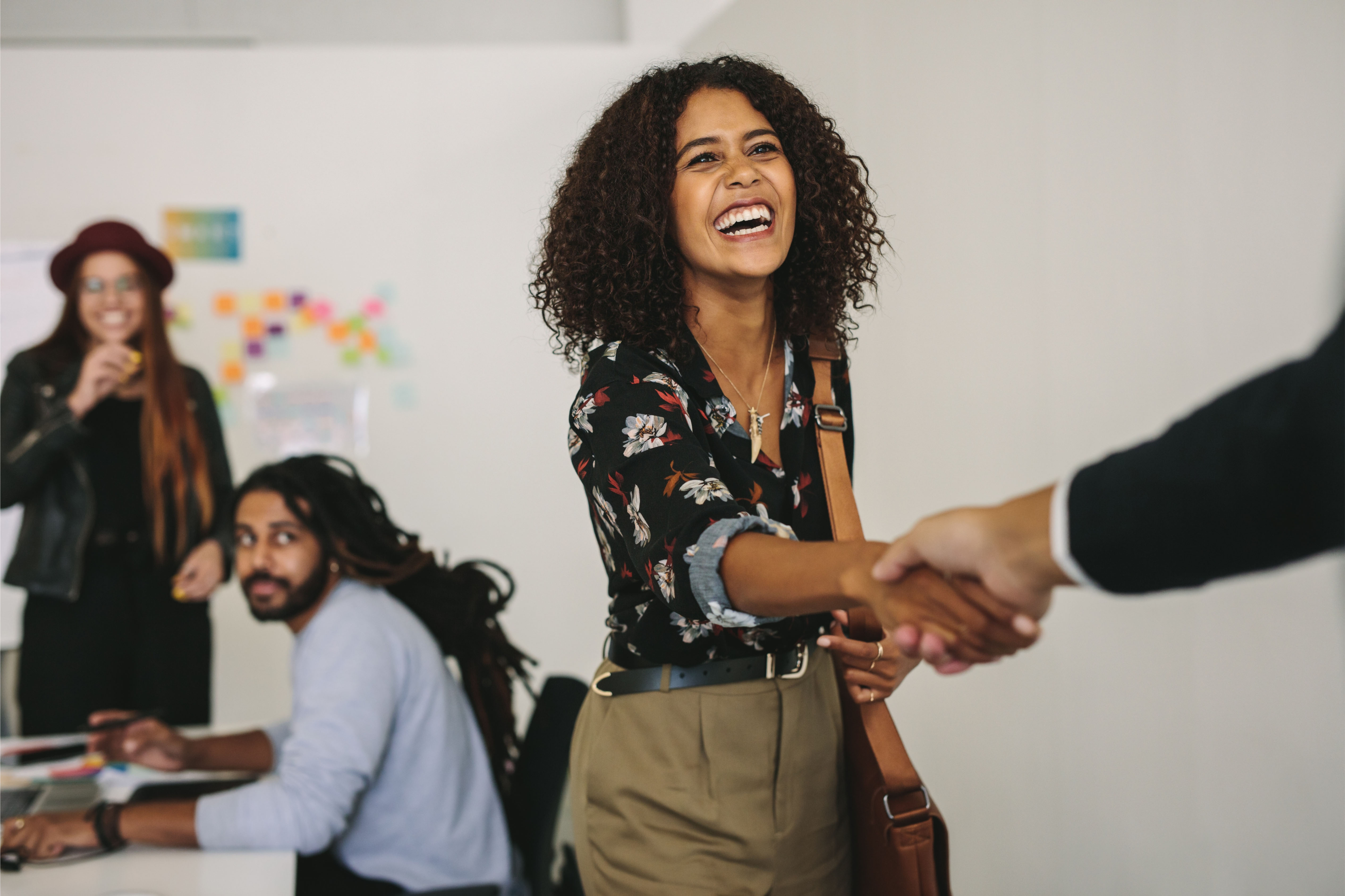 Smiling businesswoman shaking hands with a colleague in a modern office, while team members collaborate in the background.