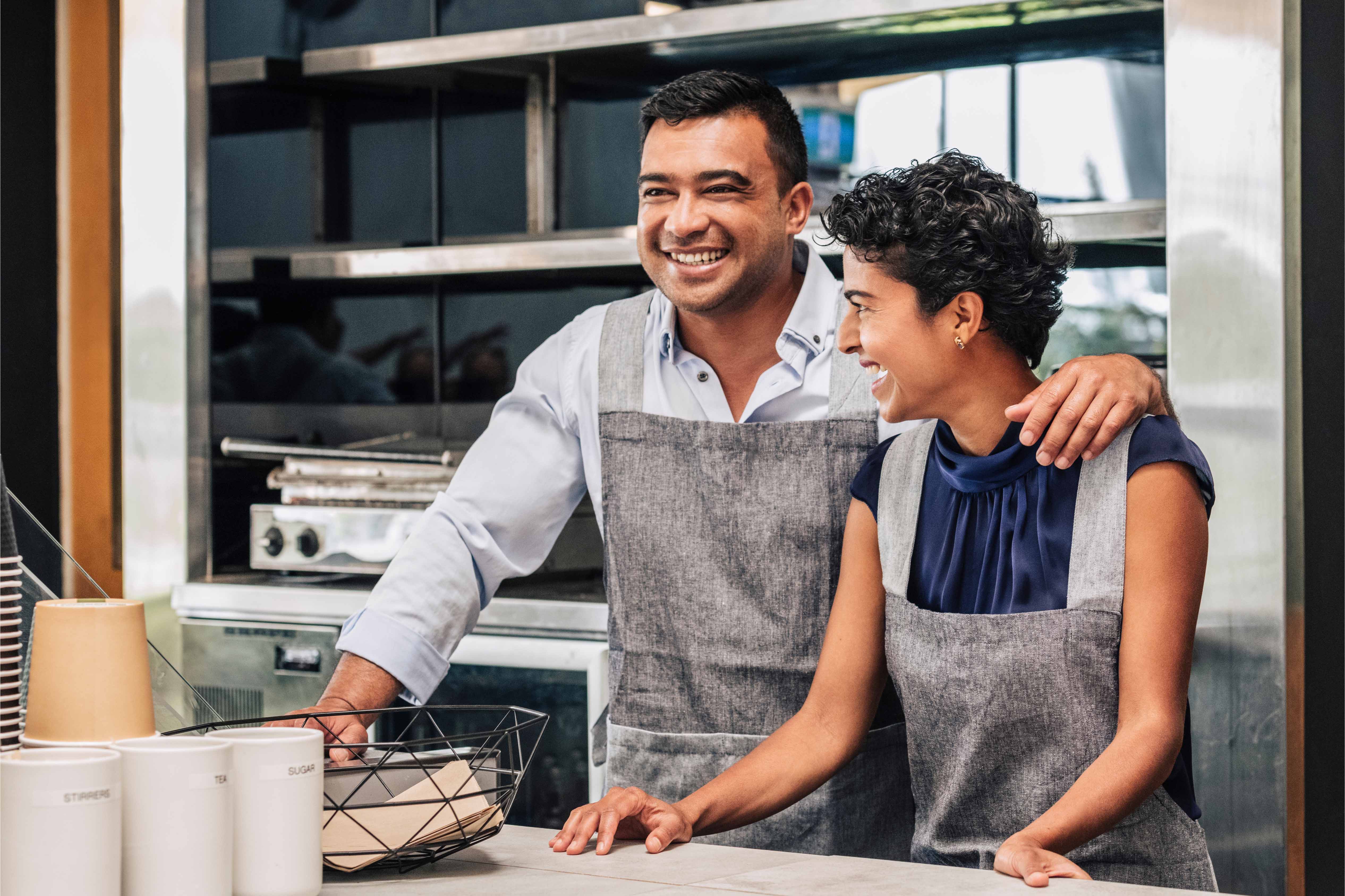 Two Mexican small business owners smiling together behind the counter of their café, wearing aprons.
