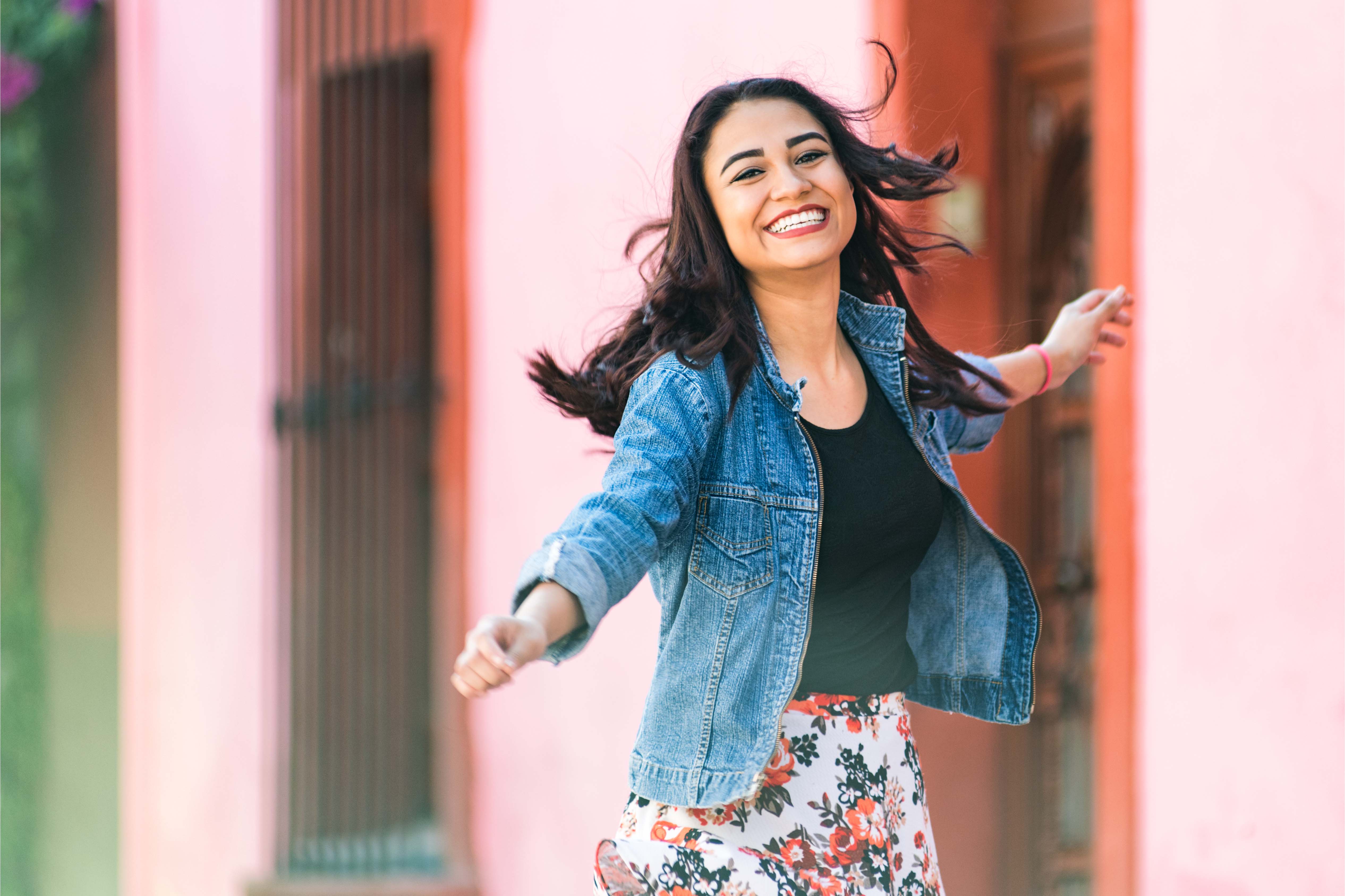 Happy young woman with long hair, wearing a denim jacket and floral skirt, smiling while walking outdoors in a colorful Mexican street.