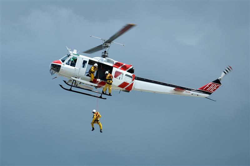 A rescue helicopter with "FIRE" on the tail hovers in the sky, while a crew member is suspended and others are aboard, wearing yellow safety gear.