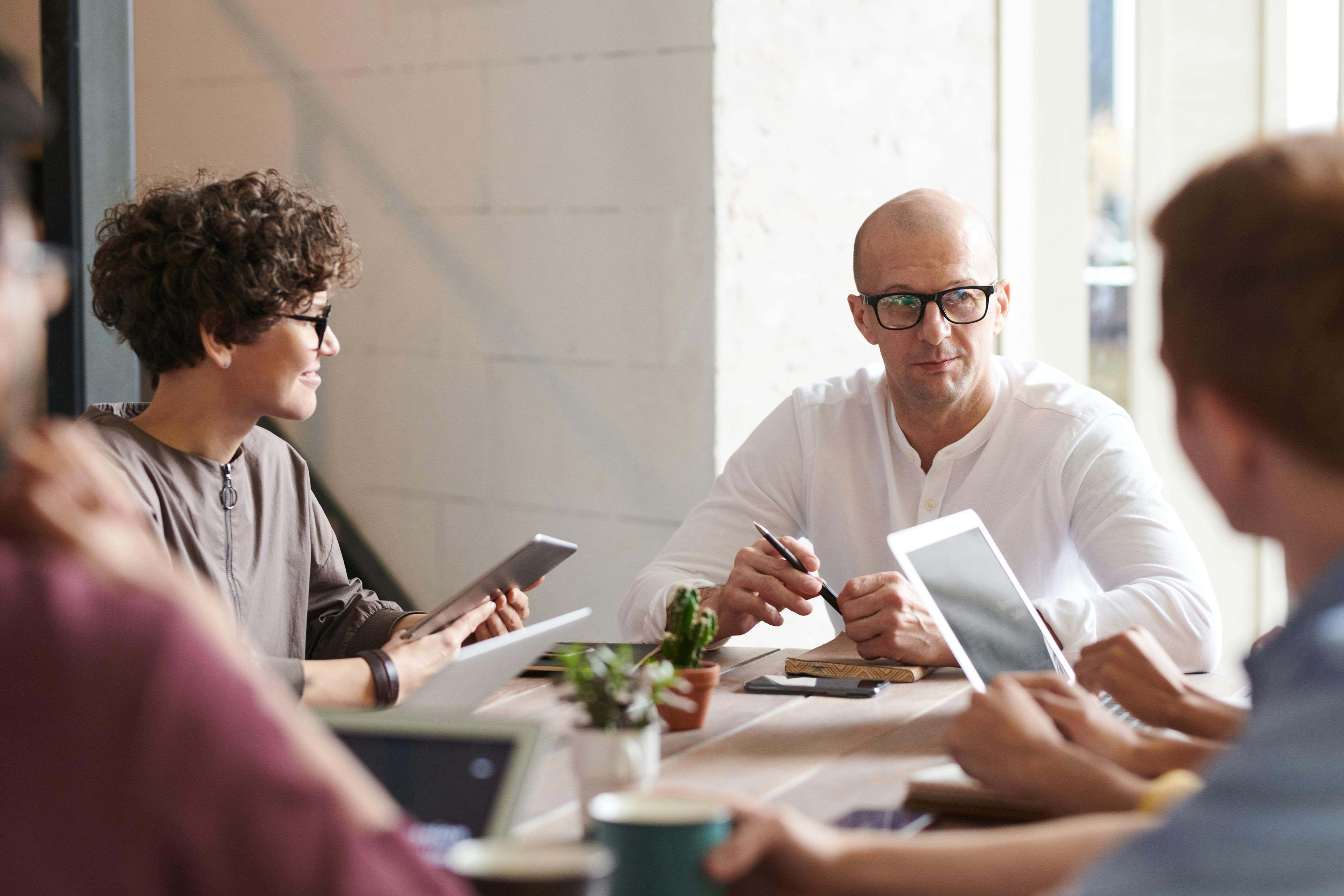 A group of coworkers sitting around a table during a meeting, with one person speaking while others listen and take notes