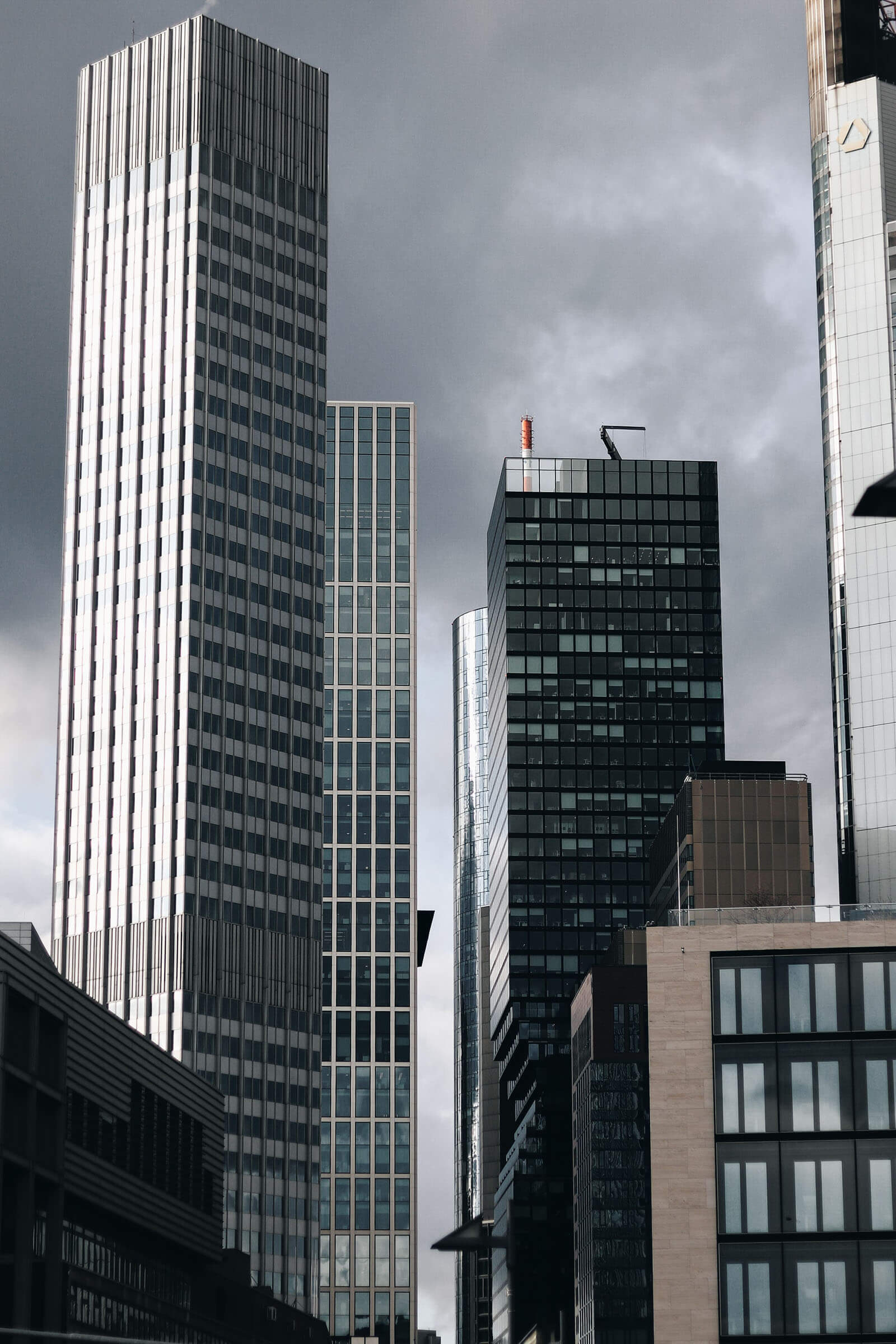 Tall modern office skyscrapers with glass and metal facades rise closely together against a dark, overcast sky.
