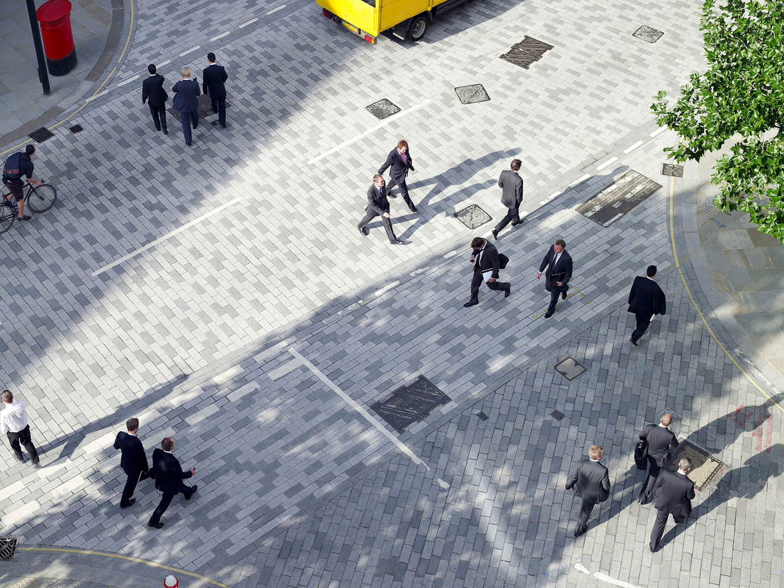 An overhead view of a wide, light-gray paved city street with multiple people in business suits walking in different directions, a cyclist on the side, a parked yellow delivery van, and tree shadows cast across the scene.