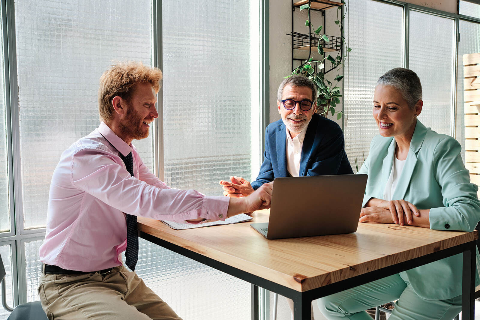 Three business professionals sit around a high wooden table in a bright modern office, smiling and collaborating as they look at a laptop screen near large frosted-glass windows and a hanging plant.