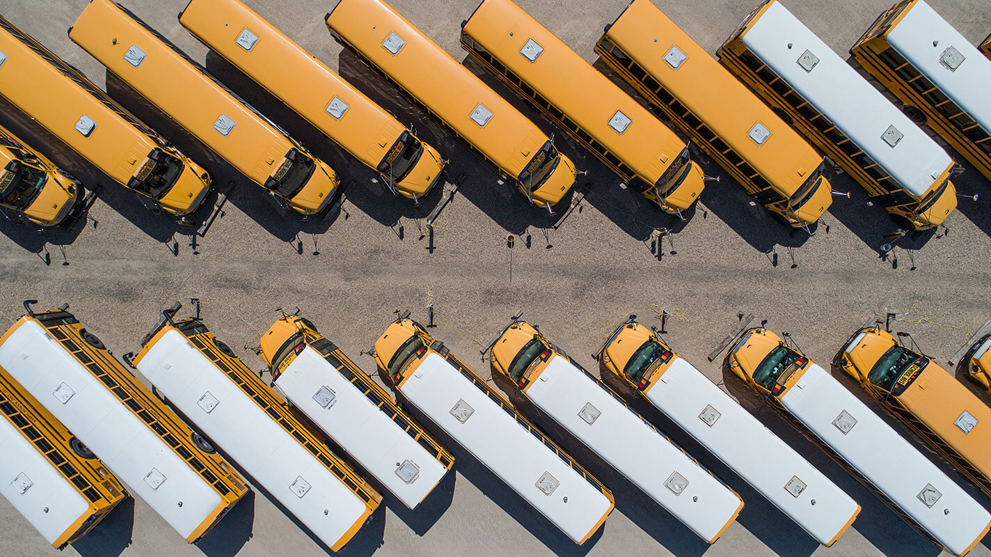 An aerial view shows multiple yellow school buses parked in neat, diagonal rows in a large lot, some with white roofs, forming a repeating geometric pattern.