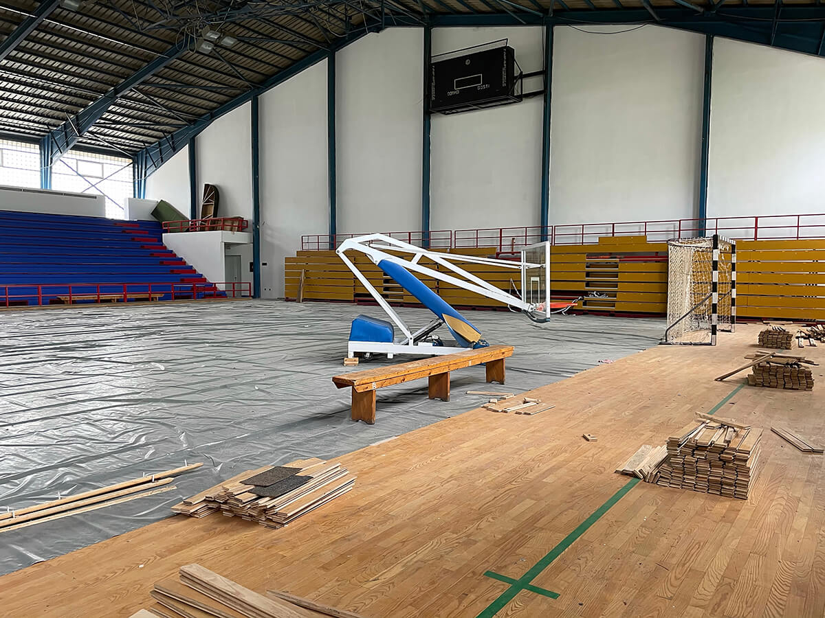 An indoor gymnasium undergoing renovation, with stacks of wooden floorboards and scattered planks in the foreground, a portable blue-and-white basketball hoop resting on a bench near the center, gray protective covering spread across most of the court, and empty tiered bleachers lining the far walls beneath a high metal roof.