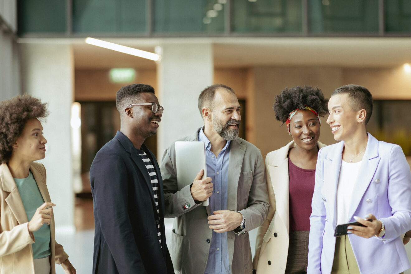 A group of five coworkers walk together through a bright modern office corridor, smiling and talking, with one holding a laptop and another a smartphone, all dressed in smart casual business attire.