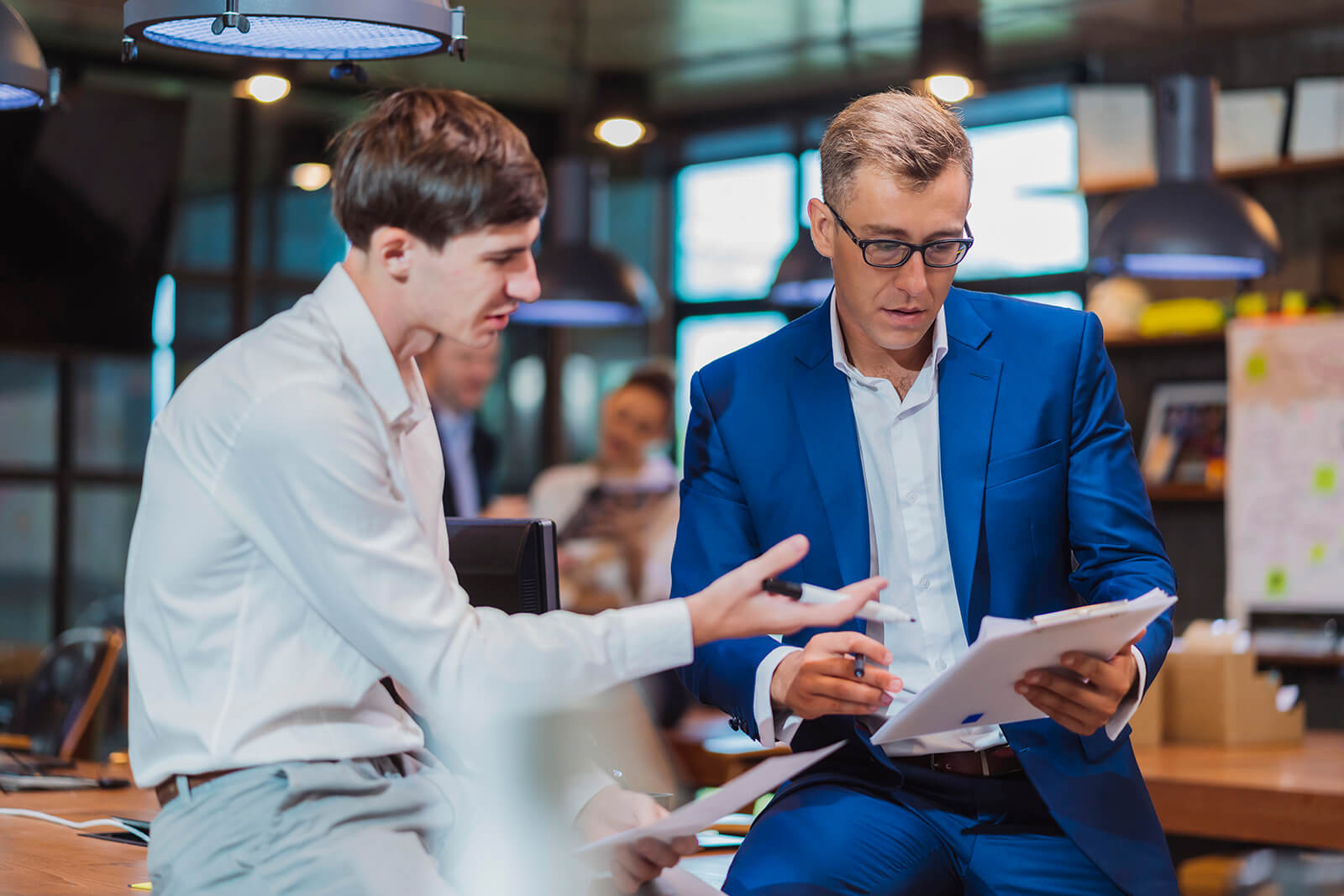 Two professionals in a modern office, one in a white shirt sitting on a desk and gesturing as they explain something, while the other, wearing a blue suit and glasses, reviews documents on a clipboard; blurred colleagues and warm pendant lighting can be seen in the background.