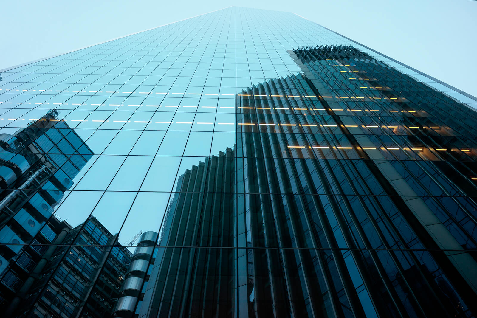 Low-angle view of a modern glass skyscraper with reflective windows mirroring nearby office buildings against a pale blue sky.